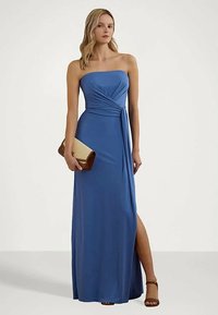 Kvinne i stroppeløs blå aftenkjole med sideslitt, holder en beige og brun clutch, iført brune ankelremsandaler.