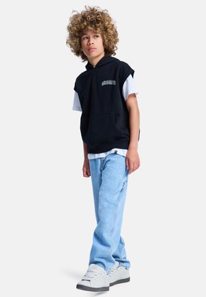 Black sleeveless hoodie with front pocket, white t-shirt underneath, light blue jeans, and white sneakers. Curly hair, standing pose.
