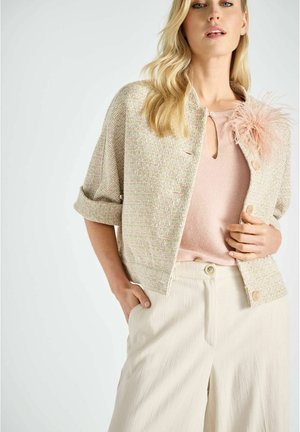 Getextureerd, crème-kleurig jasje met een cropped pasvorm, voorzien van een lichtroze veeraccent, gecombineerd met een zachtroze top en beige wijde broekspijpen.