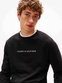 Melna sporta krekls ar balto un sarkano izšūto uzrakstu "TOMMY HILFIGER". Regulāra piegriezuma, ar apaļu apkakli un teksturētu audumu.