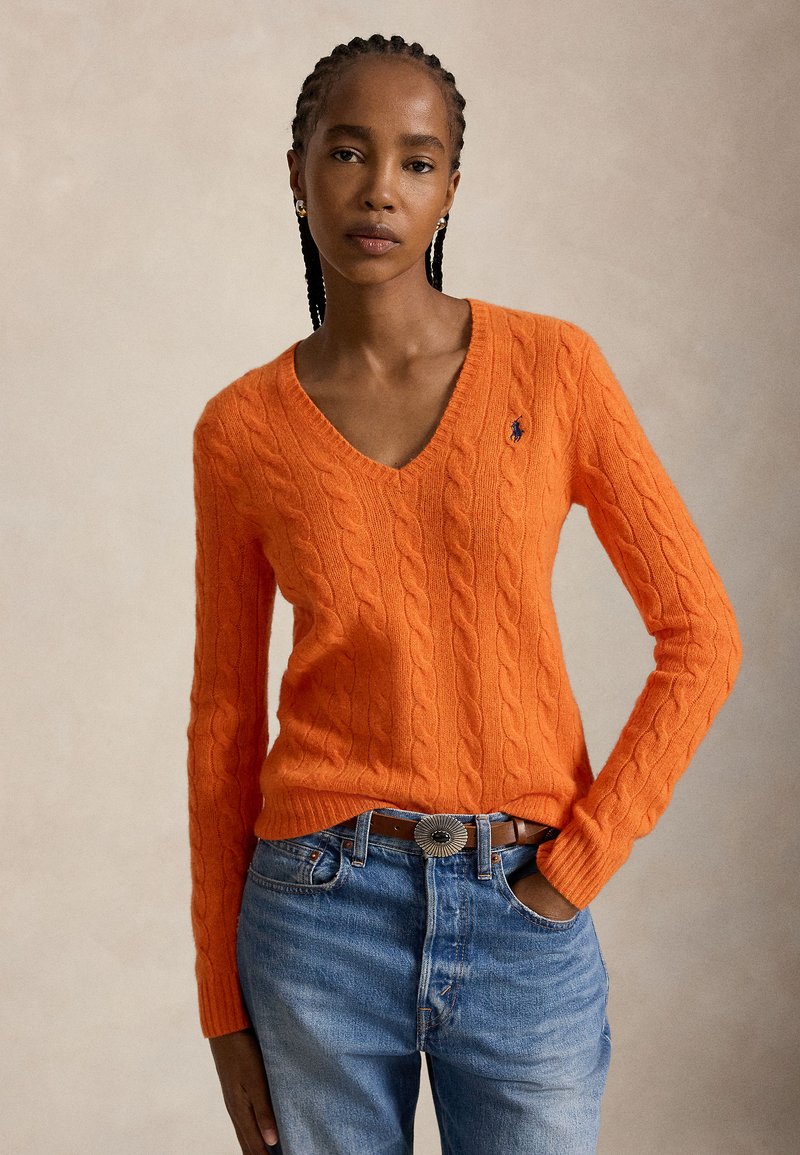 Γυναίκα με πλεξούδες, φοράει πορτοκαλί πουλόβερ με V-λαιμόκοψη πλεχτό σε σχέδιο καλωδίου και μπλε τζιν με καφέ ζώνη, στέκεται σε ουδέτερο φόντο.