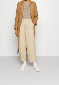 Camelmantel, beiger Strickpullover und lockere beigefarbene Hose. Weiße Sneakers, strukturiertes Material, langer Mantel mit Revers und Fronttaschen.