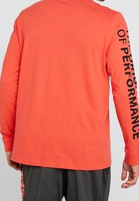 Mężczyzna ubrany w koralowy longsleeve z napisem "THE PURSUIT OF PERFORMANCE" na prawym rękawie, zestawiony z ciemnymi spodniami.