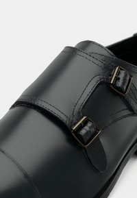 Sort læderpumps med dobbelte monk strap-spænder og fine syninger, vist i tæt optræk mod en hvid baggrund.