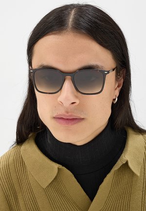 Persona con capelli lunghi e scuri indossa occhiali da sole quadrati sfumati, un dolcevita nero e un maglione verde oliva a collo alto.