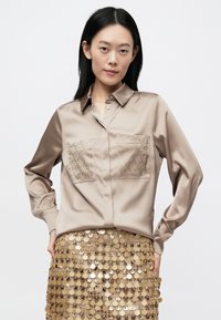 Chemise en satin beige avec deux poches à motifs, boutonnée à l'avant et manches longues, associée à une jupe scintillante en sequins dorés.