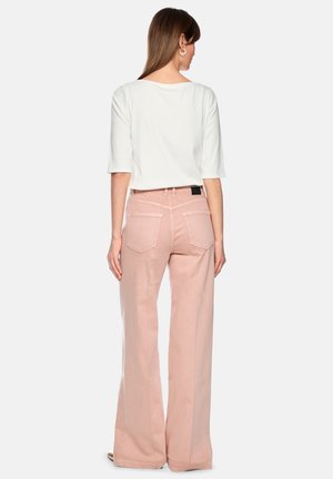 Frau trägt ein weißes Oberteil mit Ellbogenärmeln und hoch taillierte, weit ausgestellte rosa Hose, steht mit dem Rücken zum Betrachter vor einem einfarbigen Hintergrund.