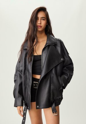 Giovane donna con lunghi capelli indossa una giacca di pelle nera oversized sopra un crop top nero e pantaloni corti con una grande fibbia, in piedi contro uno sfondo semplice.