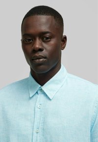 Lichtblauw linnen overhemd met een kraag, gemaakt van een gestructureerde stof en met knoopsluiting, tentoongesteld tegen een neutrale achtergrond.