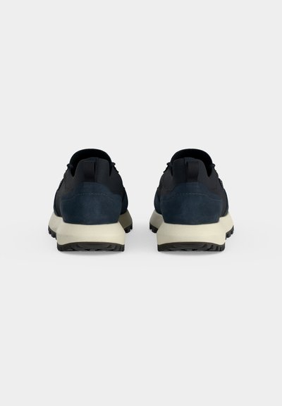Paire de baskets noires et bleu foncé avec semelles blanches vues de dos sur un fond gris clair uni.