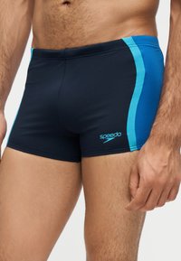 Torse masculin portant un slip de bain noir avec des bandes latérales bleues et un logo Speedo sur la cuisse gauche sur un fond uni.