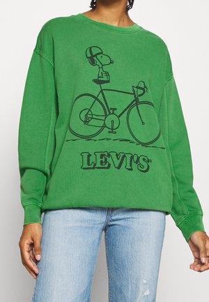 Grüner Pullover mit schwarzem Motiv von Snoopy, der einen Helm trägt und Fahrrad fährt, darunter der Schriftzug "LEVI'S", kombiniert mit hellblauen Jeans.