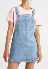 Niebieska sukienka ogrodniczka z denimu w jasnym odcieniu, z regulowanymi ramiączkami, przednią kieszenią i postrzępionym dołem. Noszona na różowej koszulce.