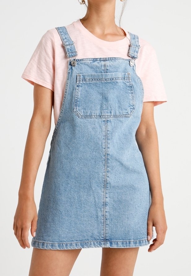 Niebieska sukienka ogrodniczka z denimu w jasnym odcieniu, z regulowanymi ramiączkami, przednią kieszenią i postrzępionym dołem. Noszona na różowej koszulce.