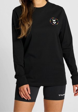 Mujer con camiseta de manga larga negra con un logotipo circular colorido y pantalones cortos deportivos negros con el logotipo blanco de "hummel" en el muslo izquierdo.