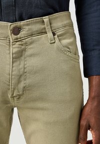 Jeans de sarga de algodón beige con un diseño ajustado, que cuentan con un pequeño bolsillo frontal, un botón de metal marrón y detalles de costura visibles.