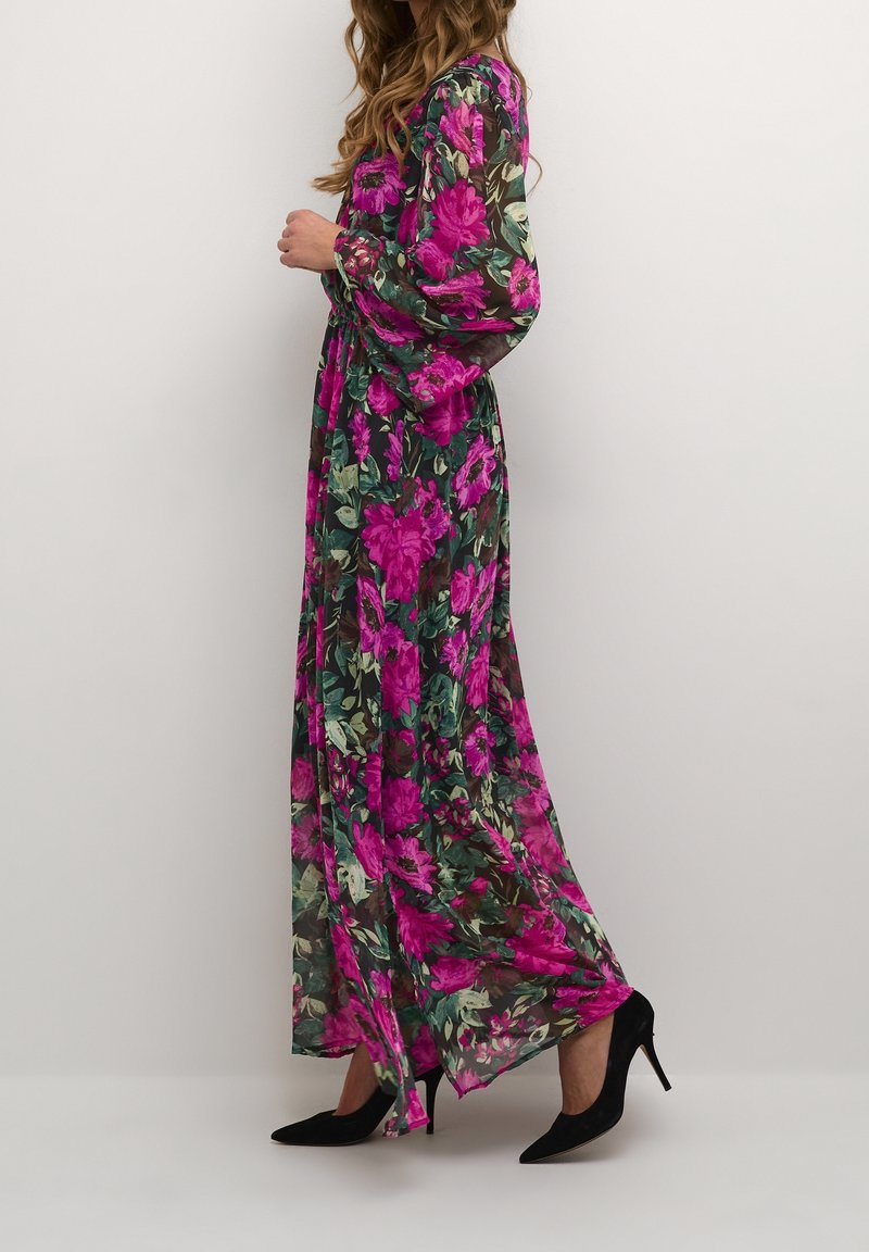 Floral Maxi-Kleid aus transparentem Stoff mit lebhaften pinken Blumen auf einem dunkelgrünen Hintergrund, langen Ärmeln und einer fließenden Silhouette. Schwarze Absatzschuhe.