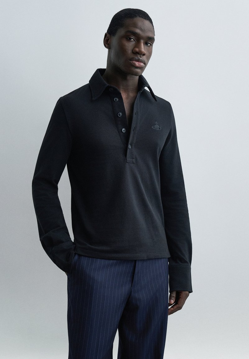 Άνδρας που στέκεται, φοράει μαύρο πόλο πουκάμισο με μακριά μανίκια και κουμπιά, καθώς και σκούρο μπλε παντελόνι με ριγέ σχεδιασμό, μπροστά σε απλό φωτεινό φόντο.