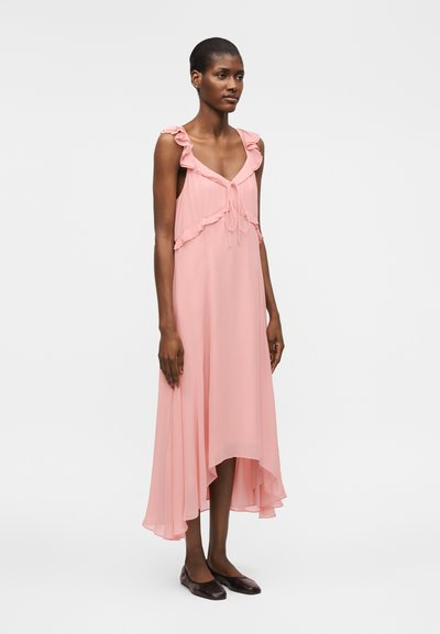 Γυναίκα με ροζ μίντι αμάνικο φόρεμα με βολάν και λεπτομέρειες με κορδέλες, σε συνδυασμό με σκούρα επίπεδα παπούτσια, στέκεται μπροστά σε απλό φόντο.