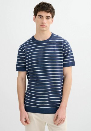 Joven con cabello oscuro y rizado, vistiendo una camisa azul marino de manga corta con rayas horizontales blancas, de pie frente a un fondo blanco liso.