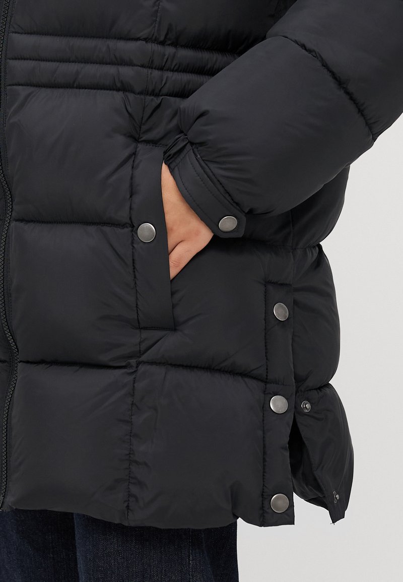 Mano en el bolsillo de una chaqueta plumón negra acolchada con botones a presión y cremallera, llevada sobre unos pantalones oscuros.