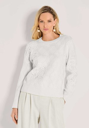 Frau in einem weißen, strukturierten Strickpullover mit floralen Mustern und kremfarbigen, weiten Hosen, die vor einem schlichten Hintergrund steht.