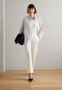 White high-waisted trousers paired with a gray and white striped shirt. Black bag held in left hand. Wooden floor and plain wall backdrop.