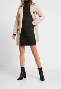 Beige trenčkot přes vypasované černé šaty s žebrovaným lemem. Černé kožené kotníkové boty s masivním podpatkem doplňují celkový vzhled.