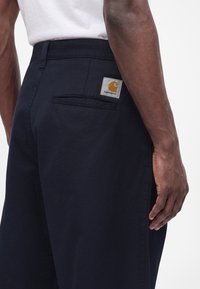 Σκούρο μπλε παντελόνι Carhartt με πίσω τσέπη και ετικέτα, φορεμένο από άτομο με σκούρο δέρμα και λευκό πουκάμισο, εμφανιζόμενο από τη μέση μέχρι το μέσο του μηρού.