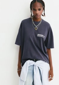 Oversized navy bomulds t-shirt med hvid grafisk print, parret med lys denim og en knyttet stribet skjorte i taljen. Sølv kædeldet.