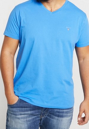 Mężczyzna w jasnoniebieskim T-shircie z dekoltem w serek i krótkimi rękawami oraz wyblakłych niebieskich dżinsach, z jedną ręką w kieszeni.