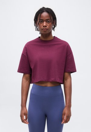 Jeune femme aux dreadlocks courtes portant un t-shirt court ample bordeaux et un legging bleu taille haute, debout devant un fond uni.