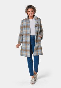 Wollmischung Mantel mit einem blauen, braunen und cremefarbenen Karomuster, ausgestattet mit einem Reverskragen und zwei vorderen Taschen, kombiniert mit blauen Jeans und Turnschuhen.