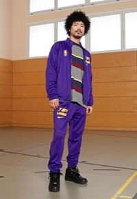 Fato de treino roxo com o logótipo amarelo dos Lakers, combinado com uma camisa cinza com riscas horizontais. Ténis de cano alto pretos completam o visual.