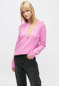 Pulover roz tricotat cu gât rotund și manșete striate, asortat cu blugi gri închis. Textură moale și croială lejeră.