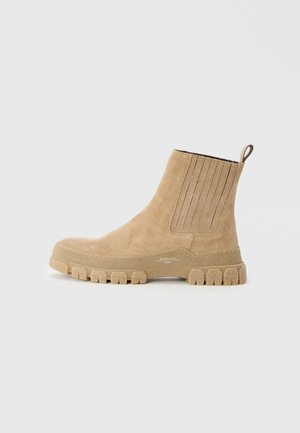 Botines de ante marrón con detalles acanalados, una lengüeta y una suela gruesa y texturizada. La vista lateral resalta la forma y el diseño estilizados.