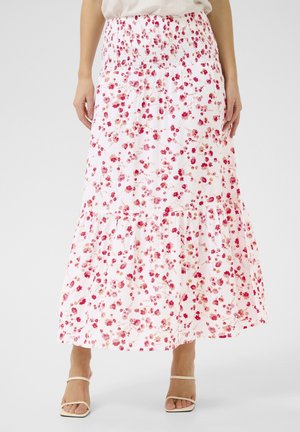 Jupe longue blanche avec imprimé floral rouge et rose, taille smockée et ourlet à volants, portée avec des sandales beiges à brides.