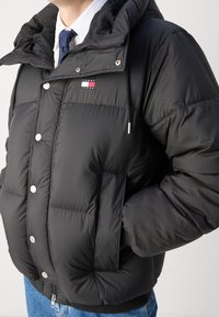 Schwarze Pufferjacke mit Kapuze, ausgestattet mit silbernen Druckknöpfen, einer Fronttasche und einem Logo-Patch. Glatte, glänzende Nylon-Oberfläche.