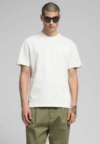 T-shirt bianca in cotone con vestibilità comoda, maniche corte e scollo rotondo, abbinata a pantaloni verde oliva. Occhiali da sole neri come accessorio.