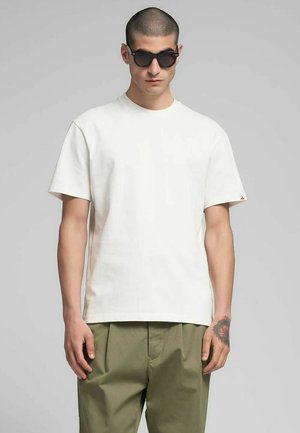 T-shirt bianca in cotone con vestibilità comoda, maniche corte e scollo rotondo, abbinata a pantaloni verde oliva. Occhiali da sole neri come accessorio.