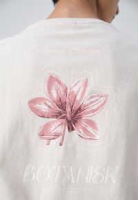 Světle šedé tričko s růžovým květinovým vzorem na zádech, se textem "Letní výstava" a "BOTANISK" pod ilustrací.