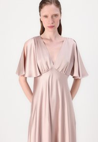 Abito in satin di un delicato rosa pallido con scollatura a V profonda e maniche a volant. Il tessuto ha una consistenza morbida e scivola dolcemente fino a un orlo al ginocchio.