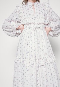 Valkoinen kukkamekko, jossa on röyhelökoristeita, pitkät puffihihat ja nauhakiristys kaulassa. Mekossa on pystysuoria raitoja ja pastellivärisiä kukkamalleja.