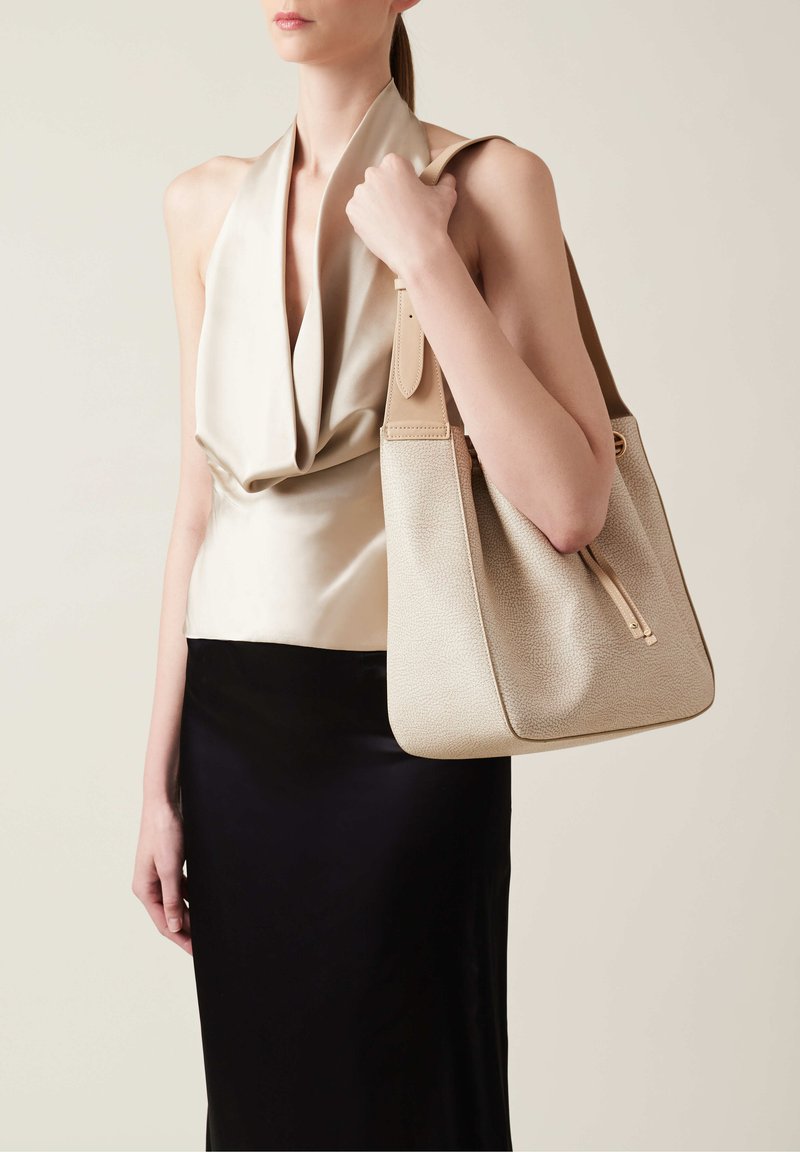 Beige leather handbag with a structured shape, pebble texture, and zipper detail, held by a person wearing a sleek, draped beige top and black skirt.