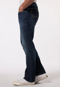 Jeans de mezclilla azul oscuro con diseño de pierna recta, que presentan una textura ligeramente deslavada y costuras amarillas contrastantes en los bolsillos. Zapatillas blancas.