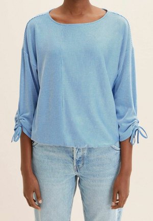 Maglietta a righe blu, dal taglio loose-fitting, con maniche a 3/4 e polsini legati. Tessuto morbido con scollatura arrotondata e orlo dritto. Indossata con jeans chiari.