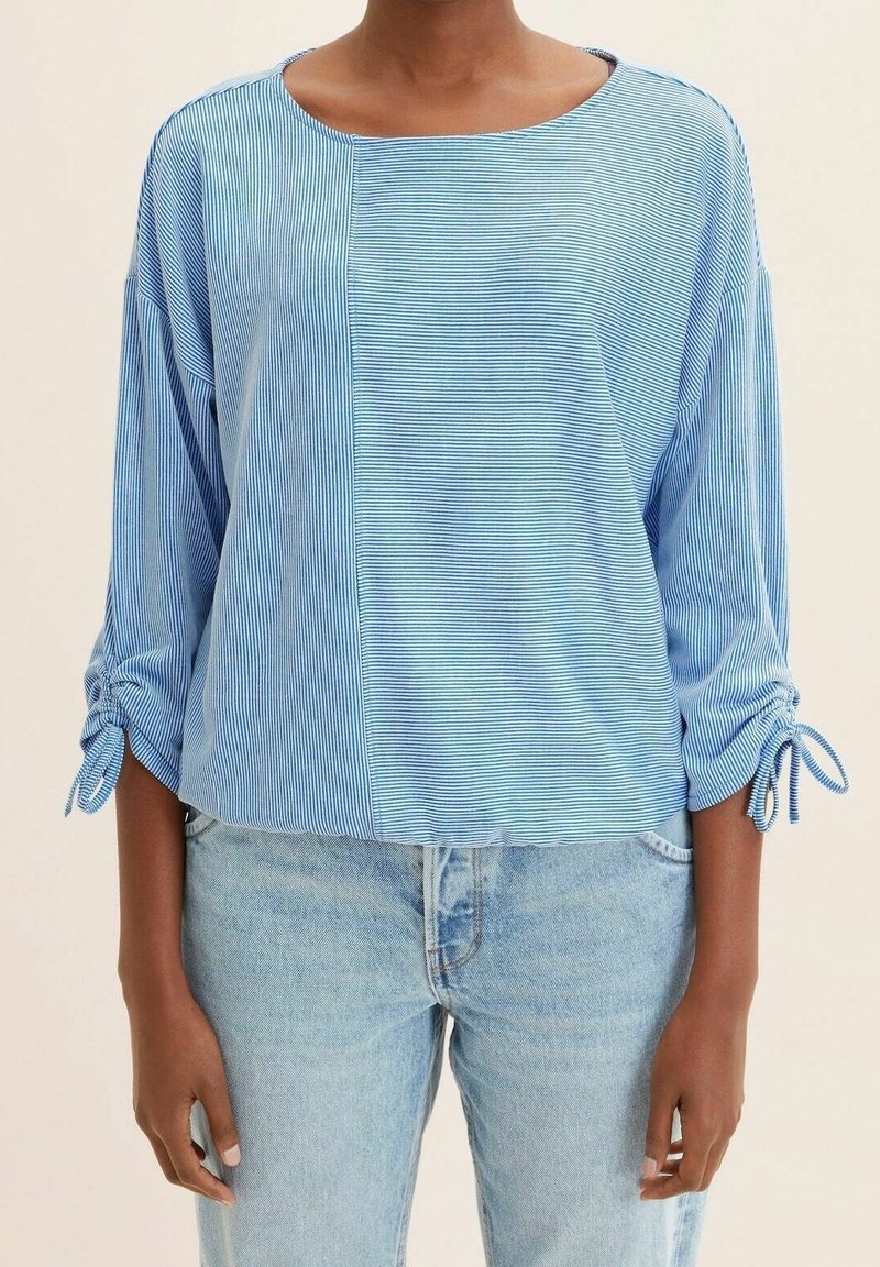 Maglietta a righe blu, dal taglio loose-fitting, con maniche a 3/4 e polsini legati. Tessuto morbido con scollatura arrotondata e orlo dritto. Indossata con jeans chiari.
