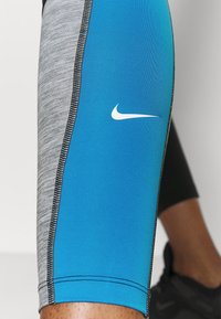 Zbliżenie nogi osoby w niebieskich i szarych legginsach sportowych Nike z czarnymi przeszyciami oraz czarnych sneakersach w tle.