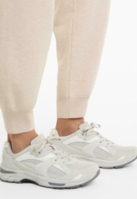 Feet wearing beige mesh and suede sneakers paired with light beige sweatpants with ribbed cuffs on a white background.