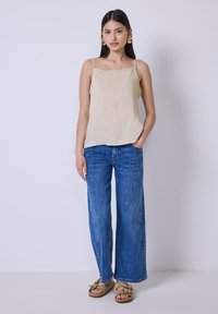 Top senza maniche color beige chiaro con scollo a onde, abbinato a jeans a gamba larga blu e sandali color cammello con hardware decorativo.